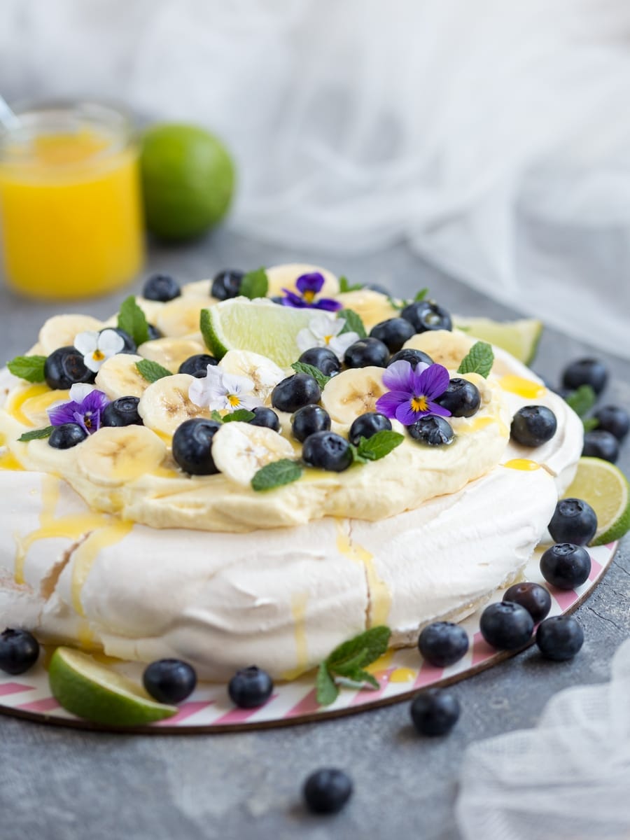Australian cake pavlova with mango cream, blueberries, banana slices, pansies, mint leaves and lime curd drizzle.