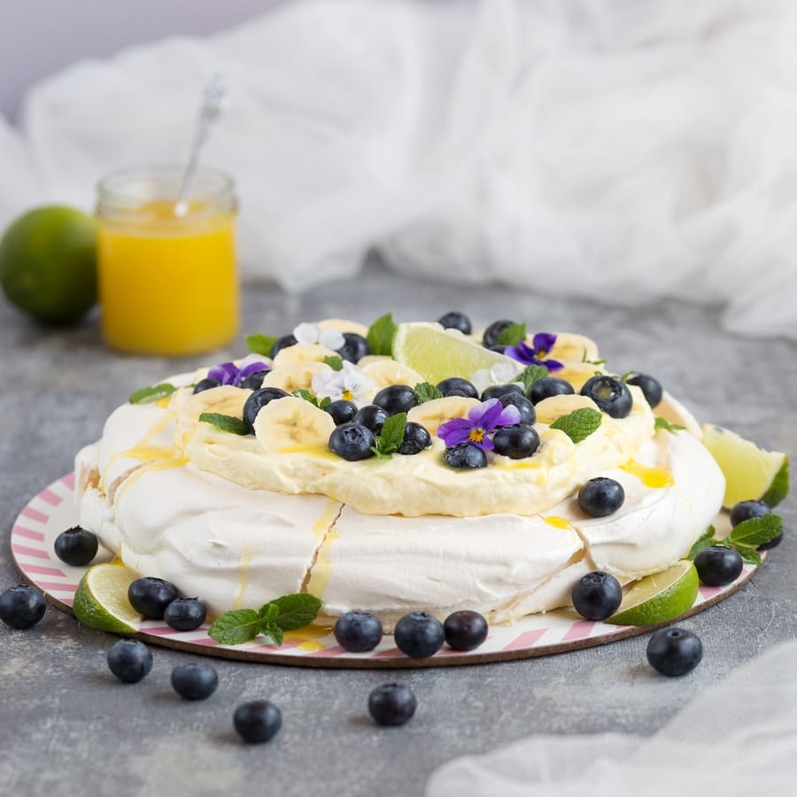 Australian cake pavlova with mango cream, blueberries, banana slices, pansies, mint leaves and lime curd drizzle.