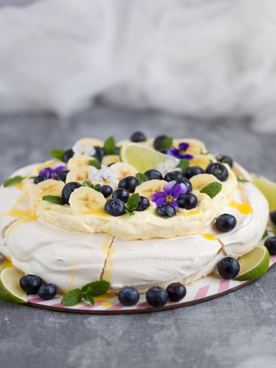 Australian cake pavlova with mango cream, blueberries, banana slices, pansies, mint leaves and lime curd drizzle.