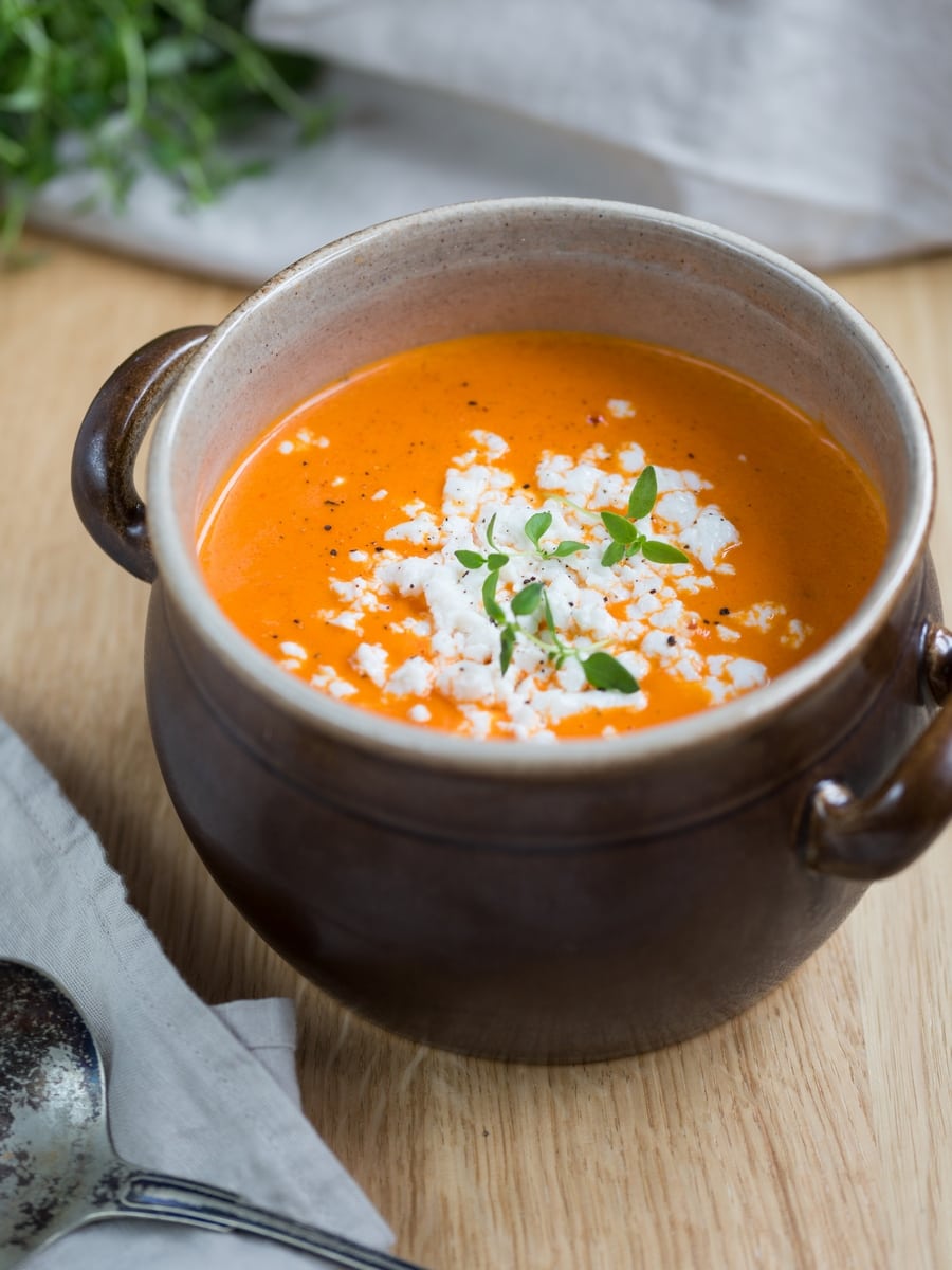 Roasted pepper soup topped with crumbed goat cheese in a brown clay pot.