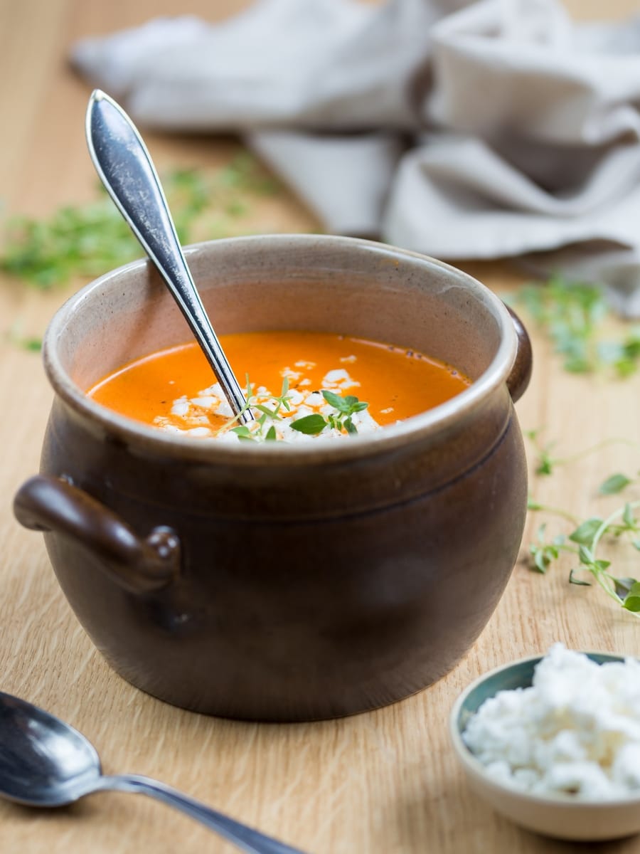 Larger view of brown clay pot with roasted pepper soup, crumbed goat cheese on top and in a bowl on the side.