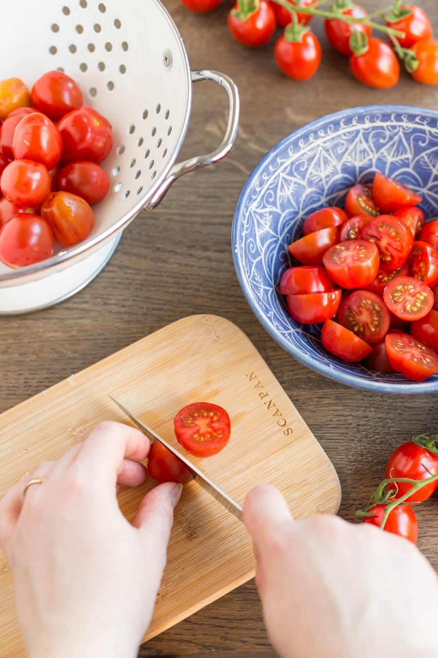 Cutting cherry tomatoes in half on a wooden board, placing cut tomatoes in a bowl.
