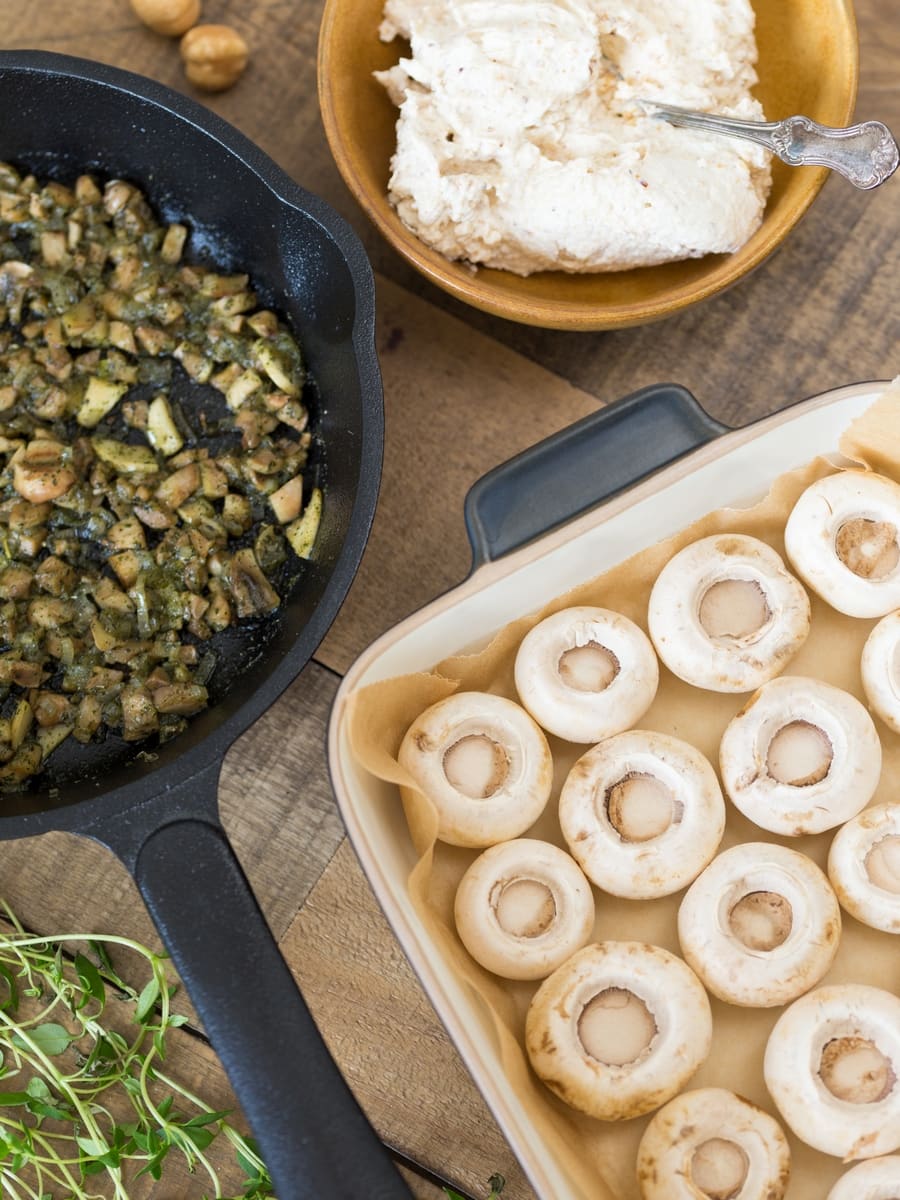 Mushroom caps upside down in an oven dish, stir fried onion and stems in a cast iron skillet, cream cheese and hazelnut mixture in a bowl.