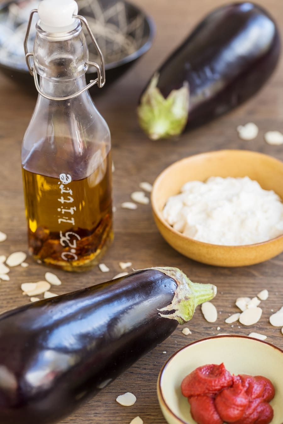 Two eggplants, tomato paste in a bowl, bottle of chili oil and ricotta in a bowl.