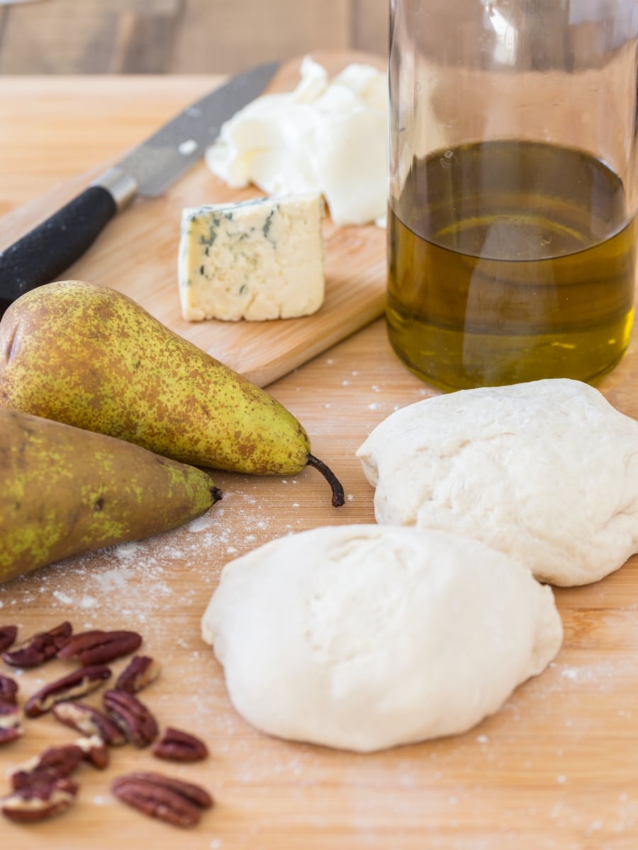 Two balls of pizza dough, chopped pecans, pears, olive oil, and blue cheese and mozzarella on a tray.