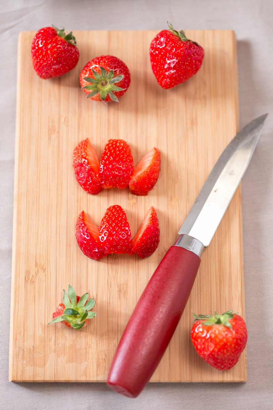 Strawberries being cut in 6 pieses with a red knife on a wooder cutting board.