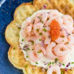 Flatlay image of waffle on blue plate topped with cream, shrimp, roe, mined onion and chives.