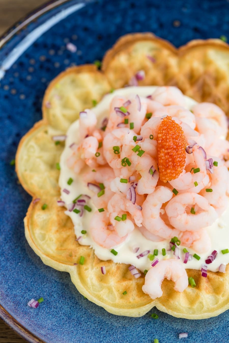 Flatlay image of waffle on blue plate topped with cream, shrimp, roe, mined onion and chives.