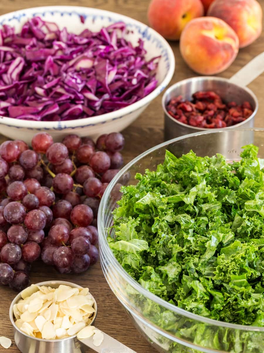 Chopped kale in a bowl, red cabbage in another, red grapes, dried cranberries in a measuring cup and shaved almonds in another.