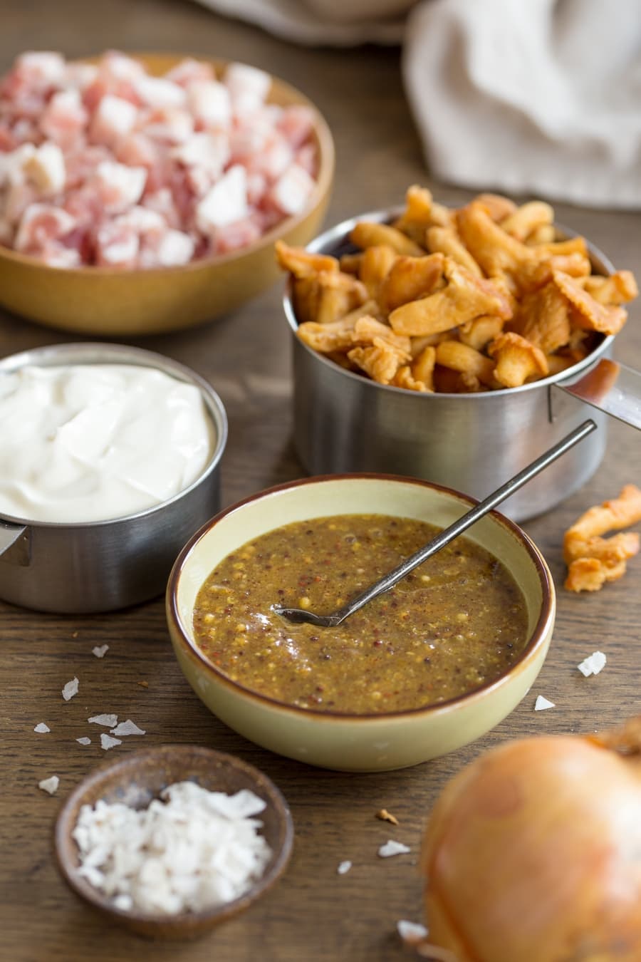 Small bowl of whole grain mustard, salt flakes and yellow onion in the foreground, cup of sour cream, chanterelle mushrooms and diced bacon in the background.