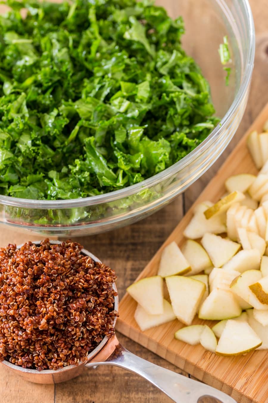 Chopped kale in a bowl, cooked quinoa in a cup and sliced pear on a chopping board.