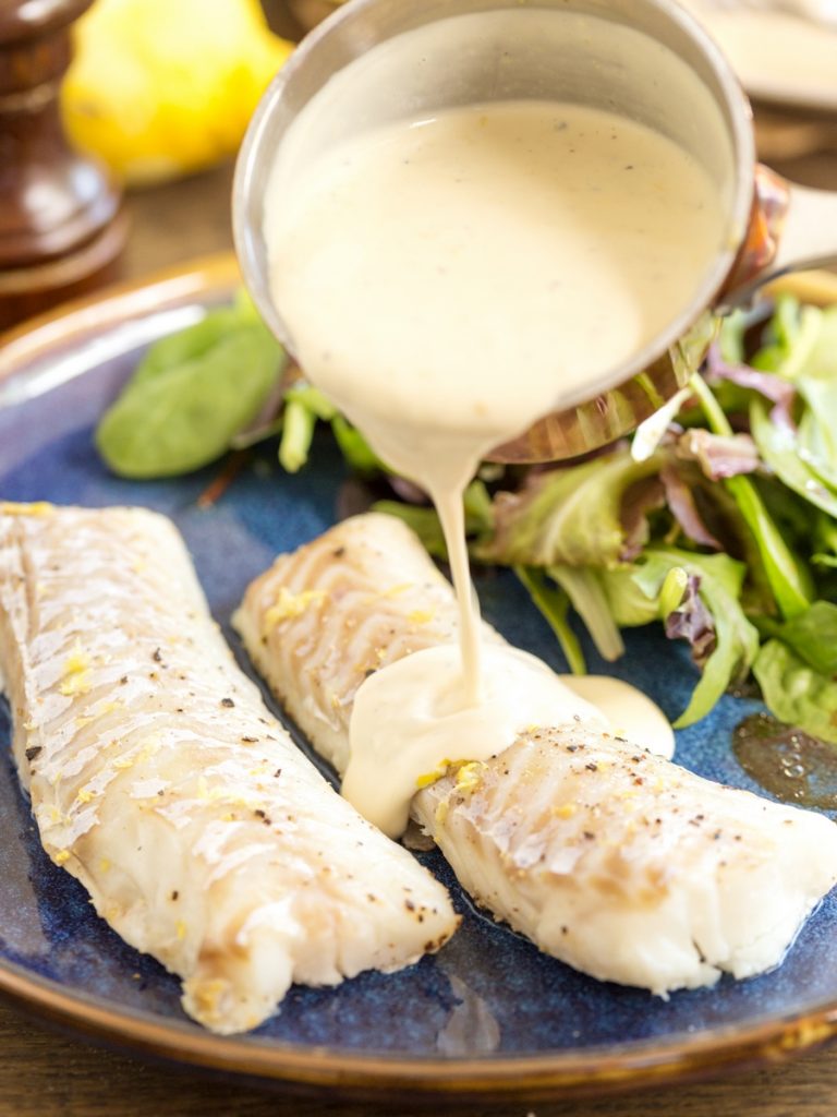 Pouring lemon pepper cream sauce over baked cod fillets.