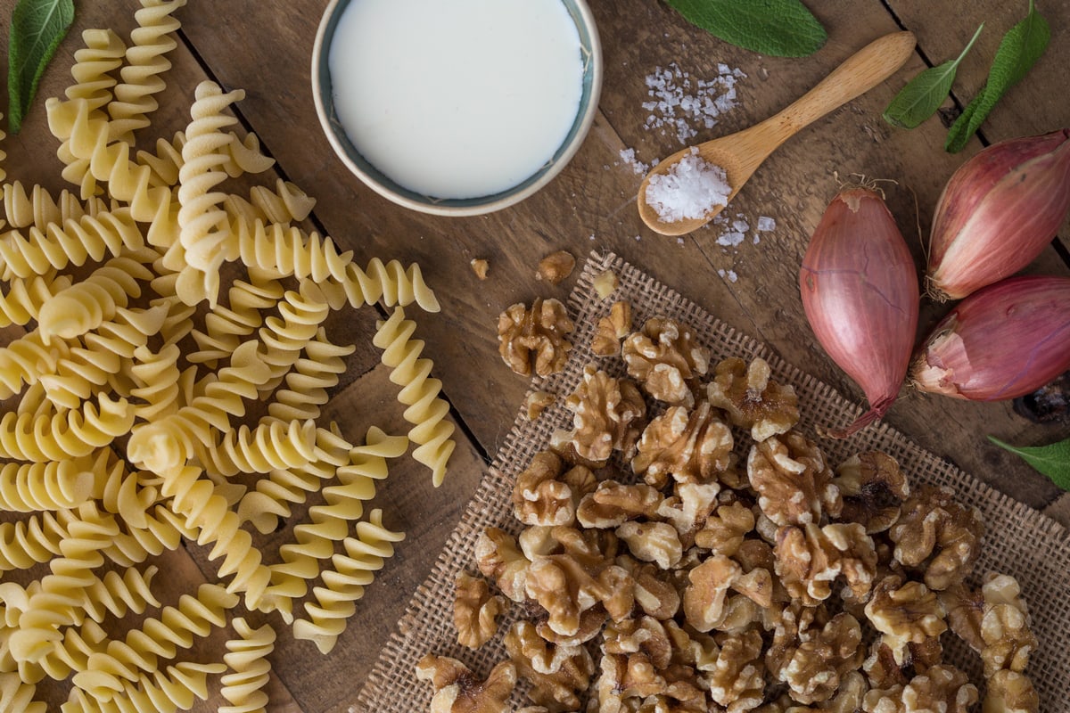 Pasta, walnuts, shallots, a spoonful of salt and a small bowl of cream on a wooden table.