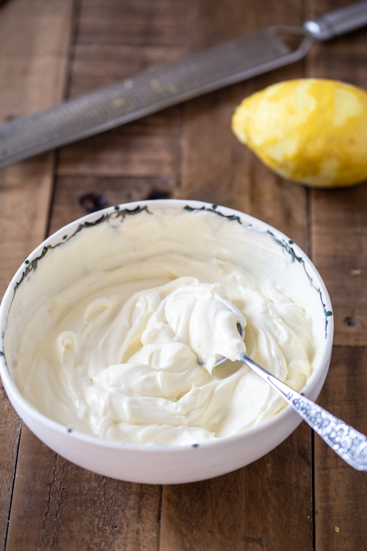 Cream cheese frosting i na bowl with a spoon inside, lemon and zester in the background.